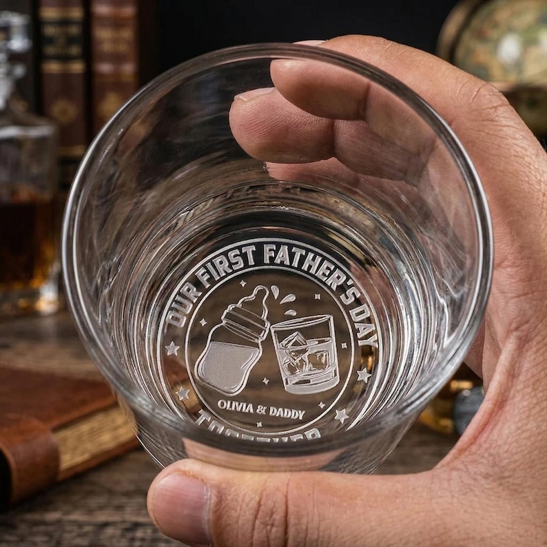 K&ouml;nnte beinhalten: Ein klares, graviertes Whiskyglas mit dem Text "OUR FIRST FATHER'S DAY" und den Namen "Olivia & Daddy". Das Design umfasst einen Babyfl&auml;schchen und ein Glas Whiskey mit Eisw&uuml;rfeln.