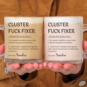 May include: Two clear glass mugs filled with a creamy beverage. Each mug has the words "CLUSTER FUCK FIXER" in bold black letters. Below the text is a definition. The name "Sandra" is written at the bottom.