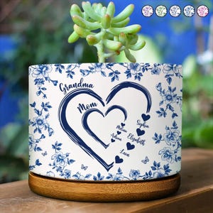 Peut inclure: Un pot de fleurs en c&eacute;ramique blanche avec un motif floral bleu et une base en bois. Le pot pr&eacute;sente un motif de c&oelig;ur avec les mots "Grand-m&egrave;re" et "Maman", ainsi que des noms. Une plante succulente pousse dans le pot.