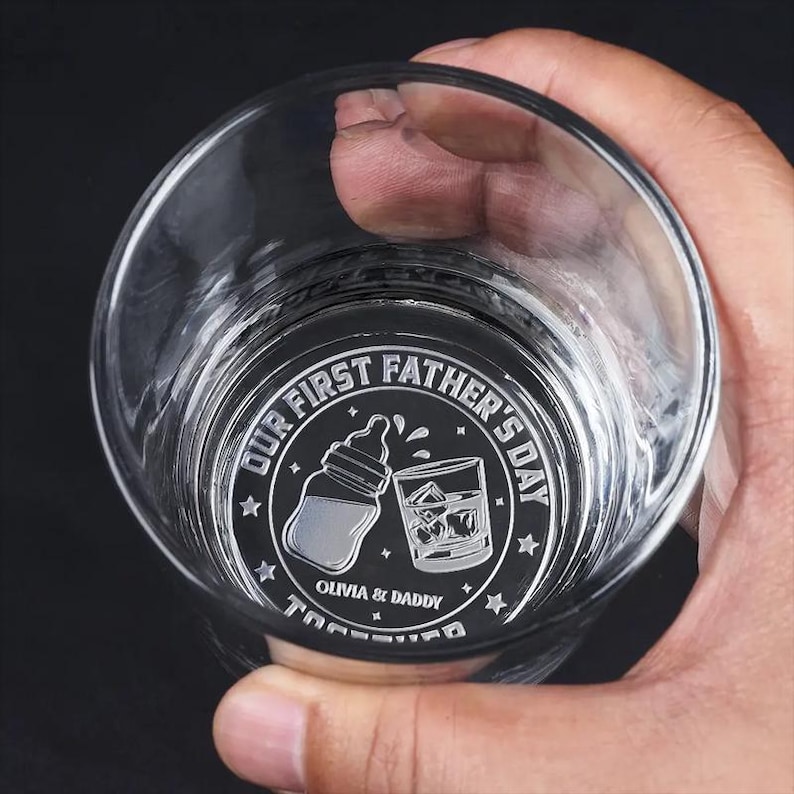 K&ouml;nnte beinhalten: Klares Glas mit kreisf&ouml;rmigem Design am Boden. Das Design zeigt einen Babyfl&auml;schchen, ein Glas Whiskey mit Eis und den Text "OUR FIRST FATHER'S DAY" und "OLIVIA & DADDY TOGETHER".