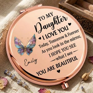 Könnte beinhalten: Ein runder, roségoldener Kompaktspiegel mit Schmetterlingsdesign und dem Text "TO MY Daughter I LOVE YOU". Der Spiegel enthält den Namen "Emily" und die Worte "YOU ARE BEAUTIFUL".