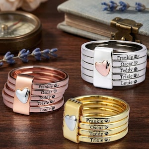 May include: Three personalized rings in silver, rose gold, and gold, each with a heart-shaped accent. The rings are engraved with names and paw prints. The rings are stacked and have a unique design.