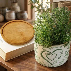 Peut inclure: Un dessous de plat rond en bois est pos&eacute; sur une pile de livres &agrave; c&ocirc;t&eacute; d'un pot d&eacute;coratif. Le pot blanc pr&eacute;sente un motif de c&oelig;ur vert avec les mots "Momma" et "Gigi" et plusieurs noms. Le pot est rempli de plantes vertes feuillues.