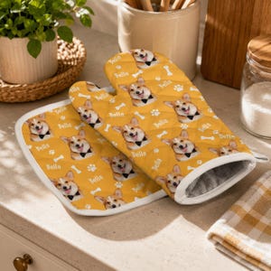 May include: A quilted oven mitt and pot holder set in mustard yellow featuring a repeating pattern of Corgi dog faces, white paw prints, white bones, and the name Bella printed in white script. The fabric has a soft, padded texture.