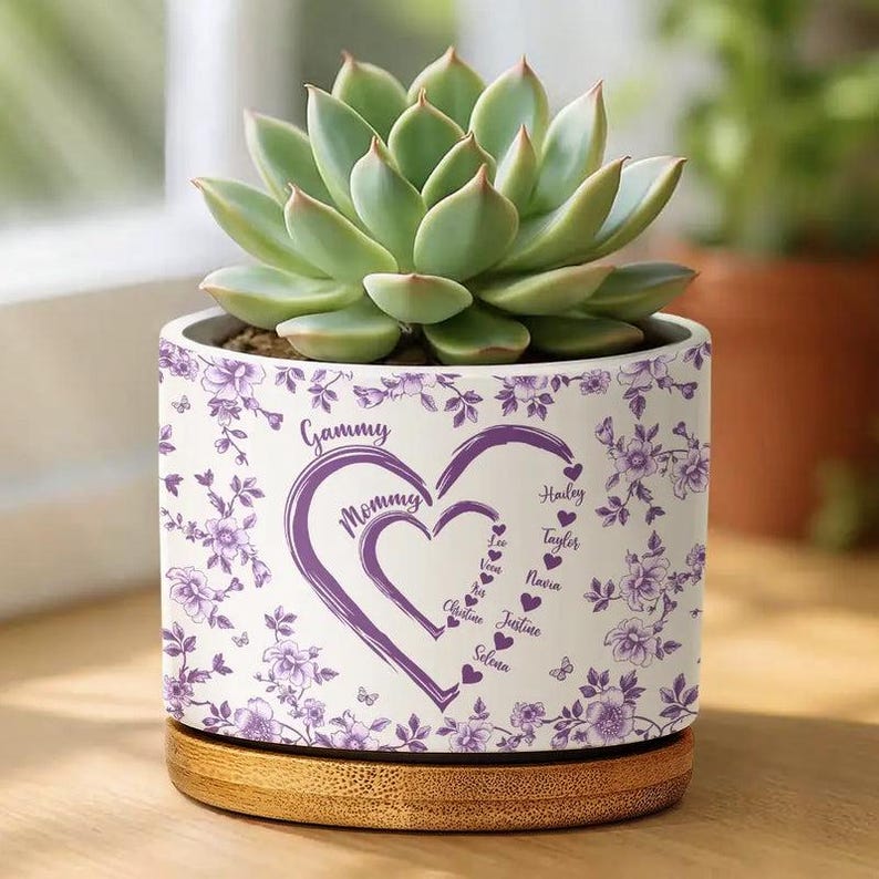 Peut inclure: Un pot de fleurs en c&eacute;ramique blanche avec une plante succulente. Le pot pr&eacute;sente un motif floral violet et un texte en forme de c&oelig;ur indiquant "Maman" et "Mamie", avec des noms en dessous. Il repose sur une base en bois.