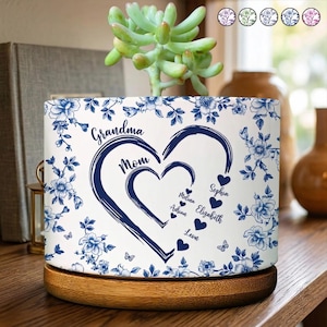 May include: A white ceramic planter with a blue floral design and a wooden base. The planter features the words "Grandma" and "Mom" with a heart design and names. A succulent plant sits on top.