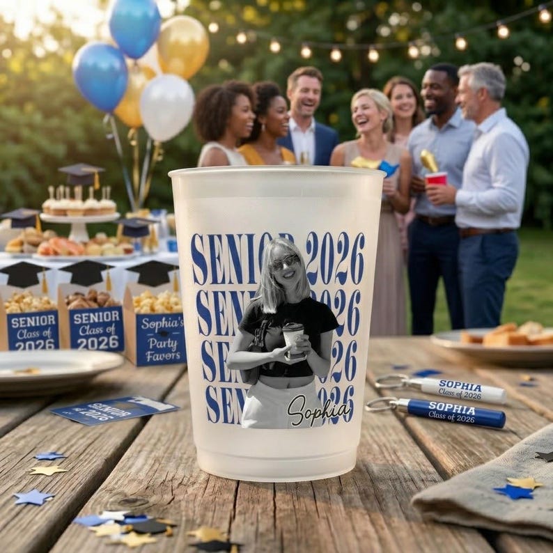 Pode incluir: Um copo de pl&aacute;stico fosco com o texto "SENIOR 2026" em azul, com uma foto de uma pessoa. O copo est&aacute; sobre uma mesa de madeira com lembrancinhas de festa e bal&otilde;es ao fundo. Chaveiros com "SOPHIA Class of 2026" tamb&eacute;m s&atilde;o vis&iacute;veis.