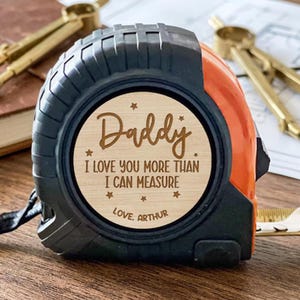 May include: Black and orange tape measure with a wooden face. The wooden face has the words "Daddy, I love you more than I can measure, Love, Arthur" engraved on it. The tape measure is on a wooden surface.