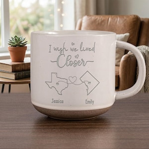 May include: A large, speckled white ceramic mug with a brown base. The mug features the text "I wish we lived Closer" and a map of Texas and Washington D.C. connected by a dotted line with hearts. The names "Jessica" and "Emily" are below the maps.