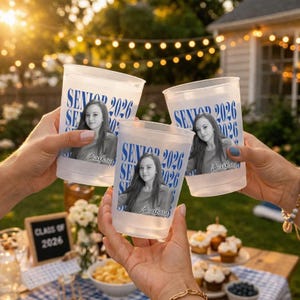 Pode incluir: Copos de pl&aacute;stico transparentes com uma foto de uma mulher, o texto azul "SENIOR 2026" e o nome "Barbara". Os copos s&atilde;o levantados em um brinde, com um fundo desfocado de uma festa no jardim.
