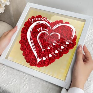 Pu&ograve; includere: Una composizione floreale a forma di cuore incorniciata. Il cuore &egrave; composto da rose rosse e presenta un testo bianco che recita "Nonna, Mamma" e nomi come "Michael" e "Sarah". La cornice &egrave; bianca e lo sfondo &egrave; giallo tenue.