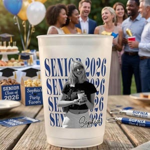Pode incluir: Um copo de pl&aacute;stico fosco com o texto "SENIOR 2026" em azul. Uma foto em preto e branco de uma pessoa est&aacute; na x&iacute;cara. O nome "Sophia" tamb&eacute;m est&aacute; na x&iacute;cara. Lembrancinhas de festa e bal&otilde;es est&atilde;o ao fundo.