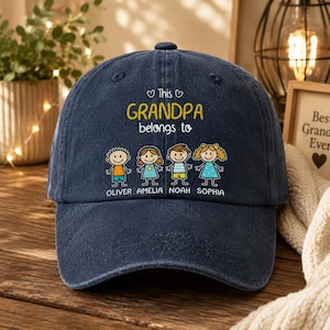 May include: A navy blue baseball cap with the text "This Grandpa belongs to" in yellow and white. Below the text are four cartoon children's figures with the names Oliver, Amelia, Noah, and Sophia. The cap is on a wooden surface.