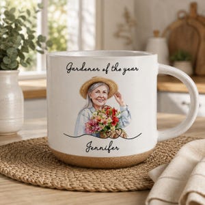 Könnte beinhalten: Ein weißer Keramikbecher mit gesprenkelter Oberfläche und beigem Boden. Auf der Vorderseite ist eine Person mit Strohhut abgebildet, die einen Korb mit bunten Blumen hält. Der Text lautet Gardener of the year und darunter steht Jennifer.
