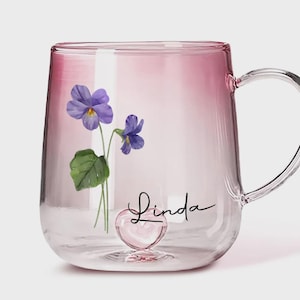 Può includere: Tazza di vetro trasparente con effetto sfumato rosa. La tazza presenta un disegno ad acquerello di viole viola e il nome "Linda" in corsivo. Una base a forma di cuore aggiunge un tocco decorativo.