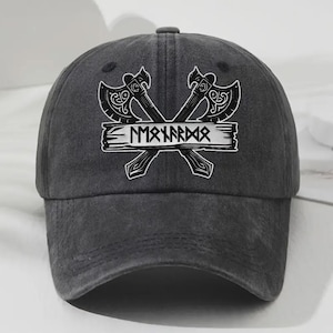 Puede incluir: Gorra de béisbol gris carbón con un gráfico blanco de dos hachas cruzadas y una pancarta con texto. Las hachas tienen detalles ornamentados. La gorra tiene una visera curva y un botón en la parte superior.