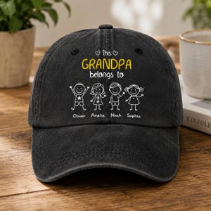 Op de afbeelding: Een zwarte baseballcap met de tekst "This Grandpa belongs to" in het geel, samen met stokfiguren en namen. De namen zijn Oliver, Amelia, Noah en Sophia. De cap is gemaakt van een zacht materiaal.