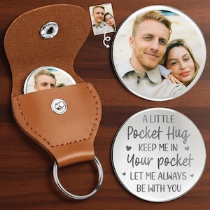 May include: A brown leather keychain with a silver ring, holding a round metal pocket hug with a photo. The pocket hug has the text "A Little Pocket Hug, Keep Me In Your Pocket, Let Me Always Be With You."