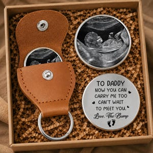 May include: A gift set comprising a brown leather keychain with a silver ring, a round metal coin with an ultrasound image, and a silver coin with the text "TO DADDY...Love, The Bump". The items are presented in a brown box.