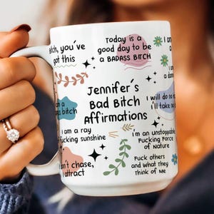 May include: White ceramic mug with the text "Jennifer's Bad Bitch Affirmations" in black, with various affirmations and decorative elements. The mug is held by a person.