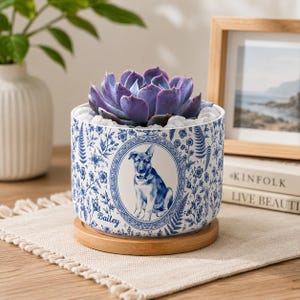 May include: A cylindrical ceramic planter featuring a blue and white floral pattern with a central oval medallion depicting a dog. The name Bailey is printed in blue script below the medallion. The planter sits on a circular wooden base.