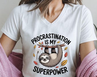 May include: White V-neck t-shirt featuring a cartoon sloth hanging from a branch. The sloth is gray and brown, with the text "Procrastination is my superpower" in black. Autumn leaves and a dandelion are also included in the design.