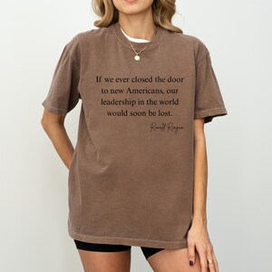 Puede incluir: Una camiseta marr&oacute;n con la cita de Ronald Reagan: "Si alguna vez cerramos la puerta a los nuevos estadounidenses, nuestro liderazgo en el mundo se perder&iacute;a pronto". La camiseta la lleva una persona.