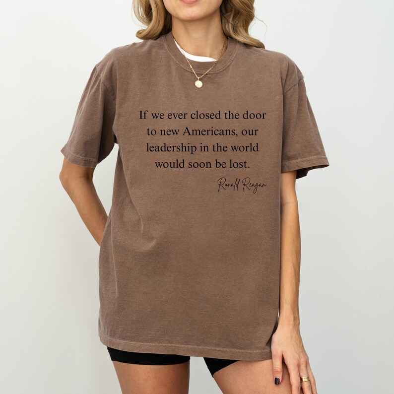 Puede incluir: Una camiseta marr&oacute;n con la cita de Ronald Reagan: "Si alguna vez cerramos la puerta a los nuevos estadounidenses, nuestro liderazgo en el mundo se perder&iacute;a pronto". La camiseta la lleva una persona.