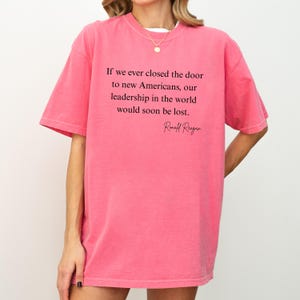 Puede incluir: Camiseta rosa con la cita de Ronald Reagan: "Si alguna vez cerramos la puerta a los nuevos estadounidenses, nuestro liderazgo en el mundo se perder&iacute;a pronto". El texto es negro. Manga corta.