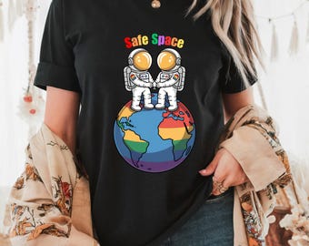 May include: Black t-shirt featuring the text "Safe Space" above a graphic of two astronauts holding hands on a rainbow Earth. The astronauts wear white suits and gold helmets. The shirt has short sleeves.