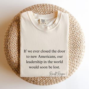 Puede incluir: Camiseta crema doblada con la cita de Ronald Reagan: "Si alguna vez cerramos la puerta a los nuevos estadounidenses, nuestro liderazgo en el mundo se perder&iacute;a pronto". Cuello redondo.