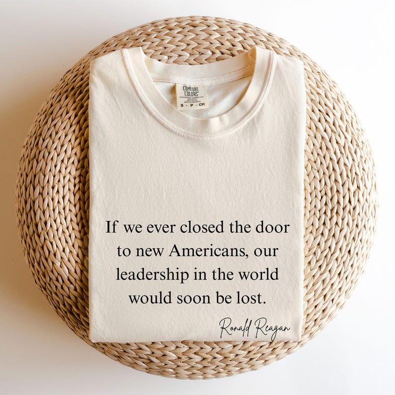 Puede incluir: Camiseta crema doblada con la cita de Ronald Reagan: "Si alguna vez cerramos la puerta a los nuevos estadounidenses, nuestro liderazgo en el mundo se perder&iacute;a pronto". Cuello redondo.
