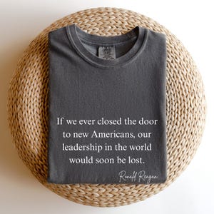 Puede incluir: Una camiseta gris oscuro con la cita de Ronald Reagan: "Si alguna vez cerramos la puerta a los nuevos estadounidenses, nuestro liderazgo en el mundo pronto se perder&iacute;a". La camiseta est&aacute; doblada sobre una superficie redonda y tejida.