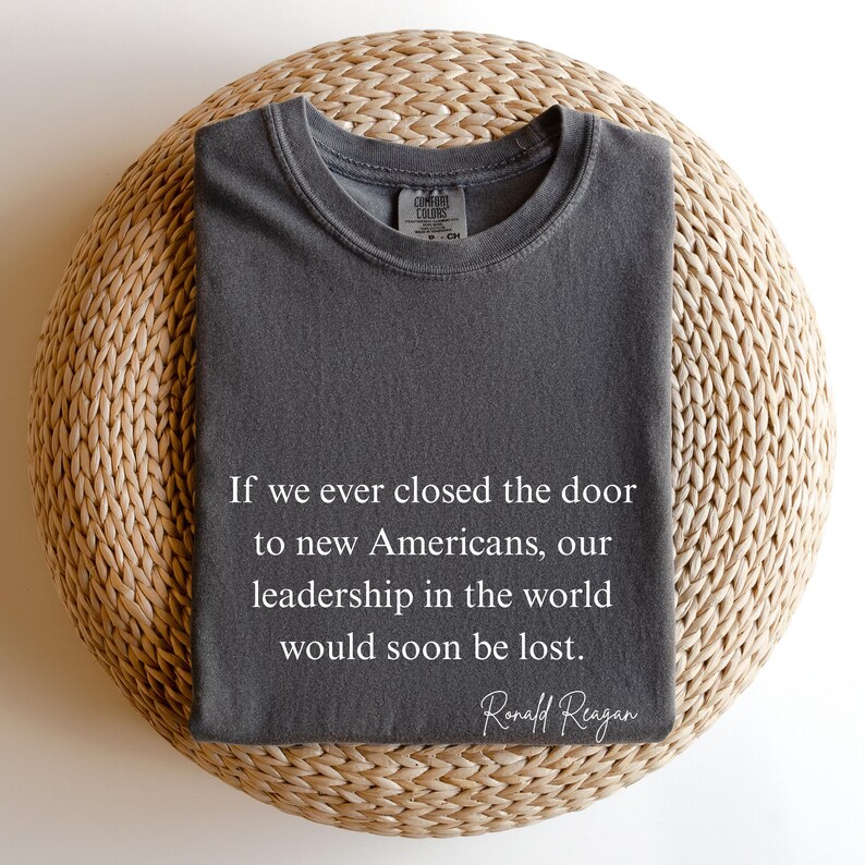 Puede incluir: Una camiseta gris oscuro con la cita de Ronald Reagan: "Si alguna vez cerramos la puerta a los nuevos estadounidenses, nuestro liderazgo en el mundo pronto se perder&iacute;a". La camiseta est&aacute; doblada sobre una superficie redonda y tejida.