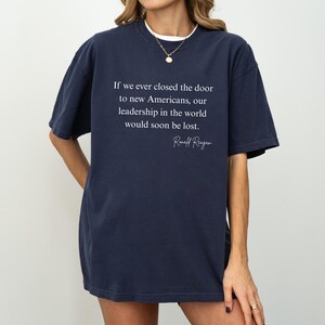 Puede incluir: Camiseta azul marino con la cita de Ronald Reagan: "Si alguna vez cerramos la puerta a los nuevos estadounidenses, nuestro liderazgo en el mundo se perder&iacute;a pronto". La camiseta tiene cuello redondo y mangas cortas.