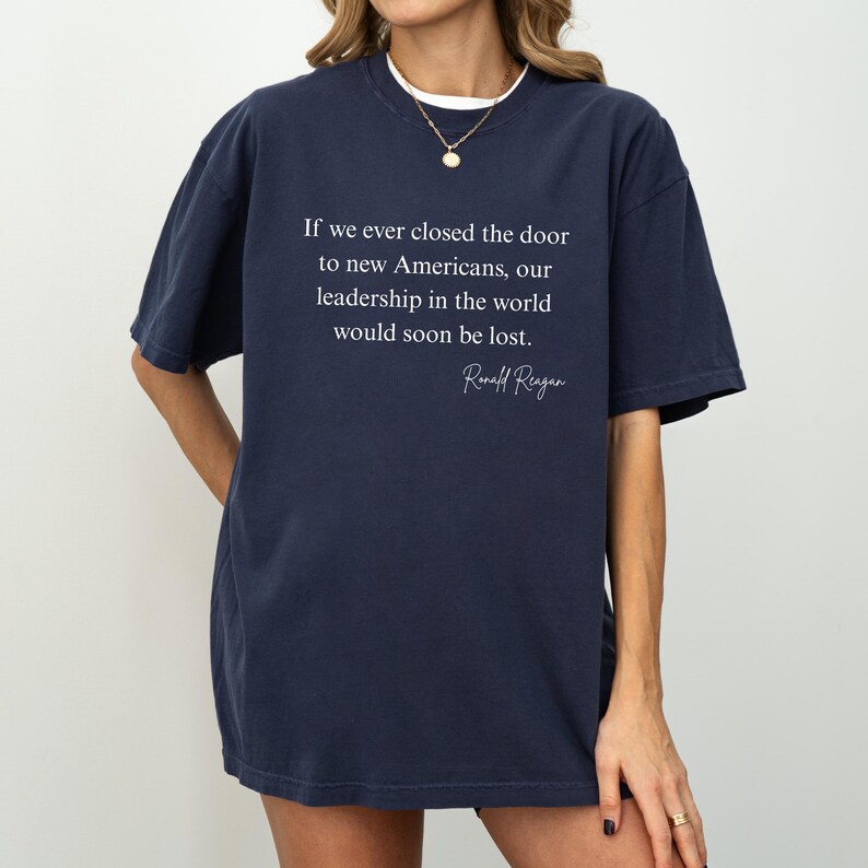 Puede incluir: Camiseta azul marino con la cita de Ronald Reagan: "Si alguna vez cerramos la puerta a los nuevos estadounidenses, nuestro liderazgo en el mundo se perder&iacute;a pronto". La camiseta tiene cuello redondo y mangas cortas.