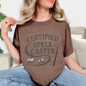 May include: A brown t-shirt with the words "CERTIFIED SPELL CASTER" in gray, along with illustrations of a wand, candles, potion bottles, and a book. The design is centered on the shirt.