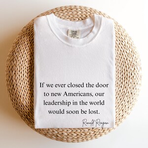 Puede incluir: Una camiseta blanca con la cita de Ronald Reagan: "Si alguna vez cerramos la puerta a los nuevos estadounidenses, nuestro liderazgo en el mundo se perder&iacute;a pronto". La camiseta est&aacute; doblada sobre una superficie redonda y tejida.