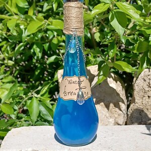 May include: A small blue glass bottle with a cork stopper and a label that reads "Water Breathing". The bottle is decorated with a silver chain and a crystal pendant.