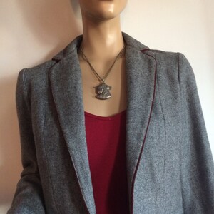 May include: A gray cropped blazer with a burgundy trim. The blazer is worn over a red tank top and blue jeans. A silver pendant necklace with a detailed design hangs from the neckline.
