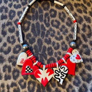 May include: A bold necklace featuring a variety of heart-shaped pendants in red, white, and black. The necklace has a mix of beads and a silver-colored clasp. The hearts have unique patterns, including a checkerboard and abstract designs.