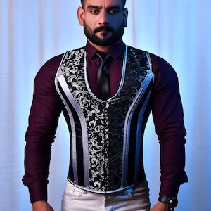 May include: A man wearing a black and silver patterned vest with a burgundy shirt and white pants. The vest has a V-neck and a black tie.