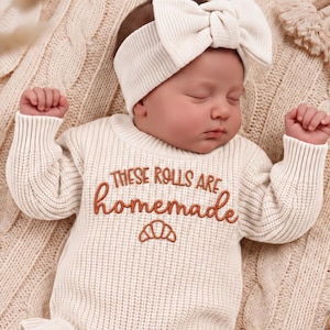 May include: A cream-coloured long-sleeve knit romper with a ruffled hem and matching headband and socks. The romper features the embroidered text 'THESE ROLLS ARE homemade' in brown with a small croissant icon centred below the lettering.