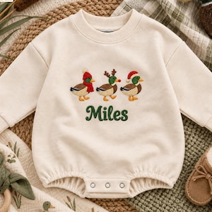 May include: A cream-coloured long-sleeve baby romper featuring an embroidered design of three mallard ducks wearing winter accessories, including a red hat, reindeer antlers, and a Santa hat, with the name Miles embroidered in green below the ducks.