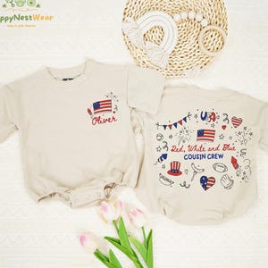 May include: Two beige baby rompers with patriotic designs. One features an American flag and the name "Oliver", the other has "USA", "Red, White and Blue", and "Cousin Crew" with festive illustrations. A woven placemat and flowers are in the background.