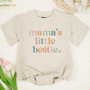May include: A beige baby romper with the text "mama's little bestie" in pastel colors. The short-sleeved romper has a round neckline and snap closures at the bottom. The romper is made of a soft material.