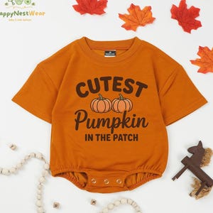 May include: Orange short-sleeved baby bodysuit with the text "Cutest Pumpkin in the Patch" and two pumpkin illustrations. The bodysuit has snap closures at the bottom and is surrounded by fall-themed decorations.