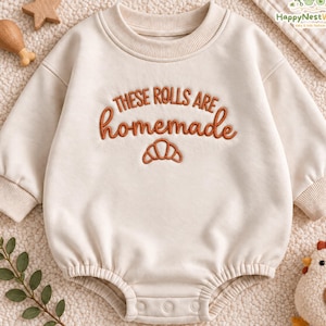 May include: A cream-coloured long-sleeve romper featuring the embroidered text "THESE ROLLS ARE homemade" in rust-coloured thread, accompanied by a small croissant icon. The garment has a ribbed crew neckline and cuffs with snap closures.