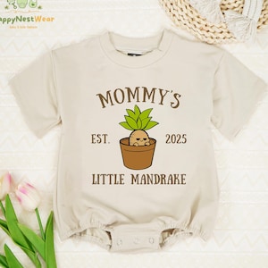 May include: Beige baby onesie with the text "MOMMY'S EST. 2025 LITTLE MANDRAKE" and a cartoon mandrake plant illustration. The onesie has short sleeves and a snap closure. The onesie is made of a soft material.