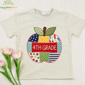 May include: A cream-colored short-sleeve t-shirt featuring a large, multi-patterned apple appliqué on the front. The apple design includes various fabrics with stars, polka dots, stripes, and pencil prints, with the text 4TH GRADE stitched in white.